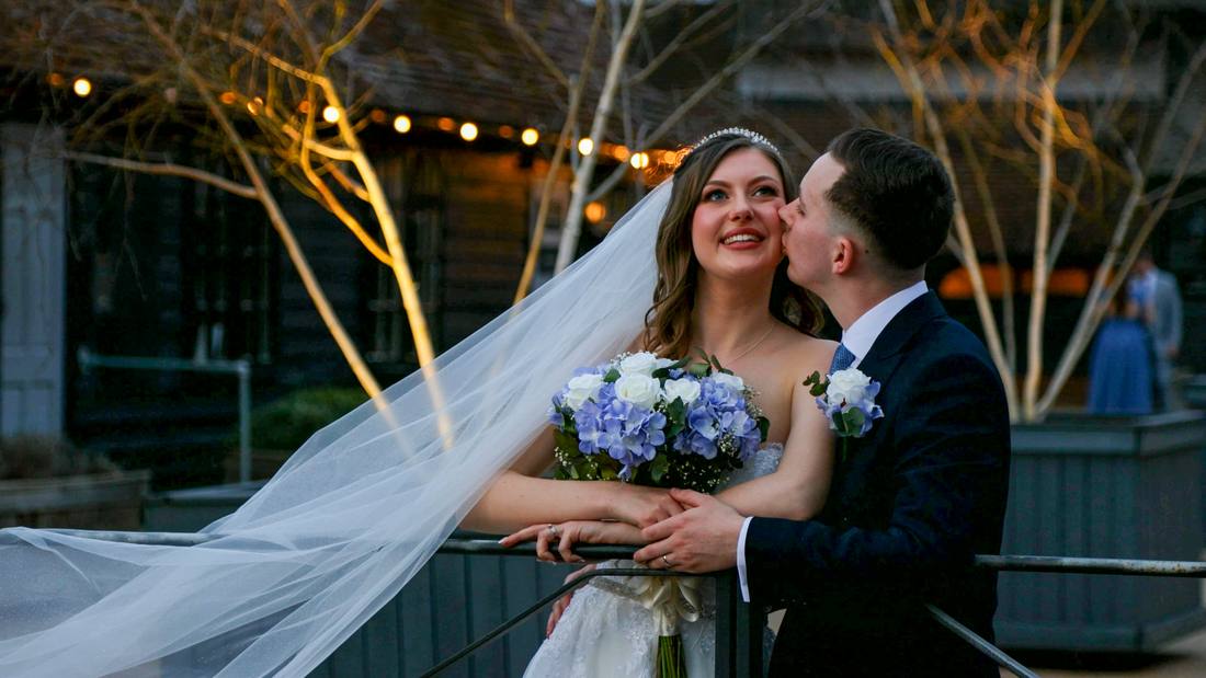 A groom kissing his bride's cheek, her veil caught in the wind