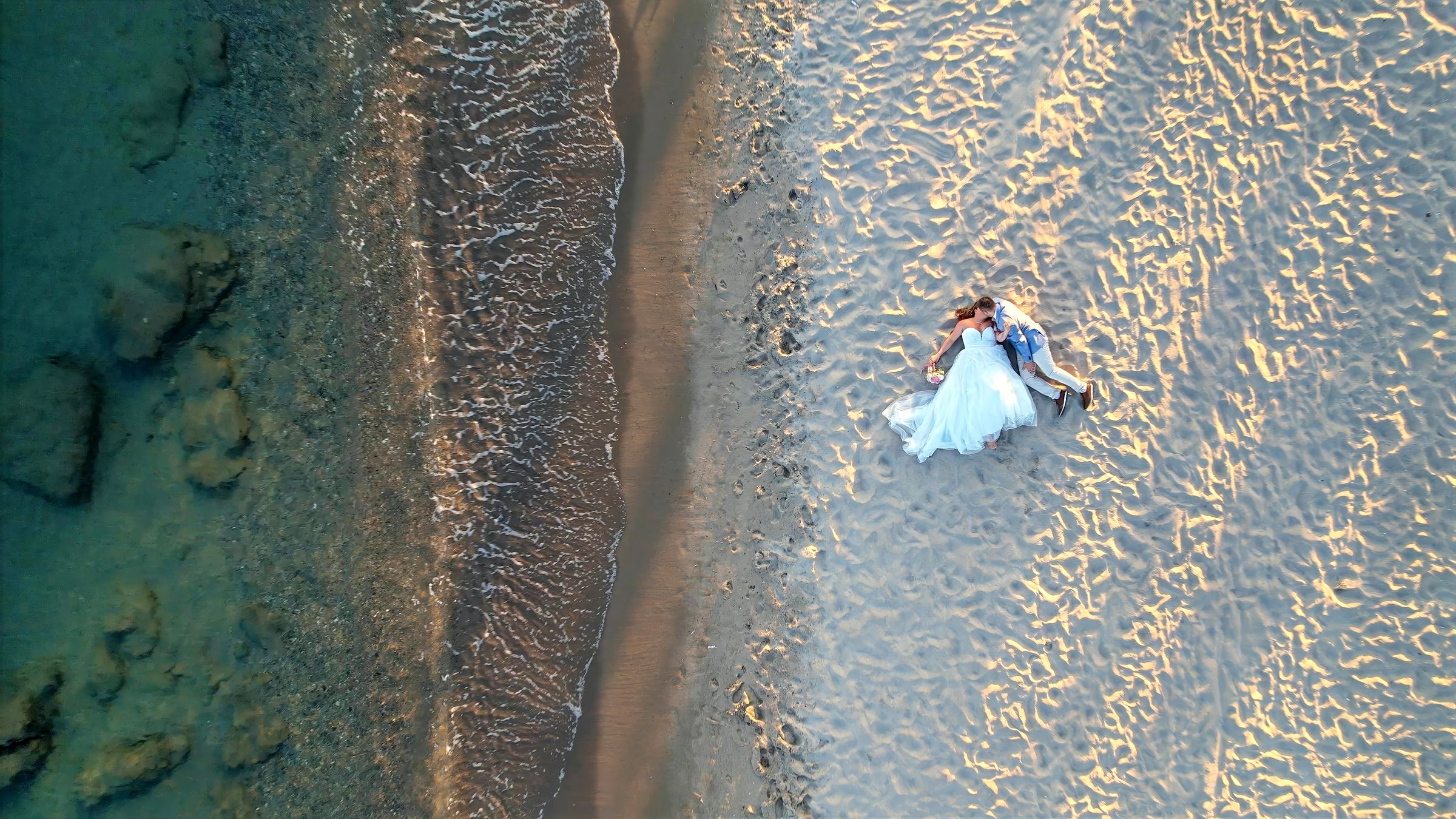 Aerial view of a bride and groom lying together on a beach where the sea meets the sand