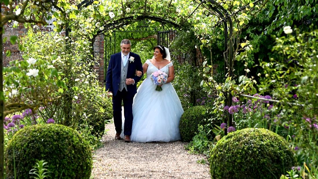 A bride walking through a rose garden arch with her father