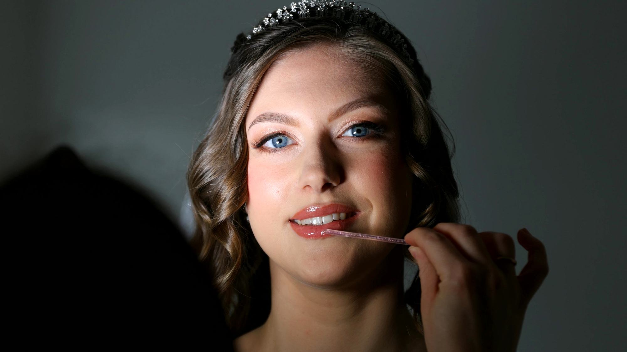 A bride in a tiara having her make-up finished, lit softly against a dark wall